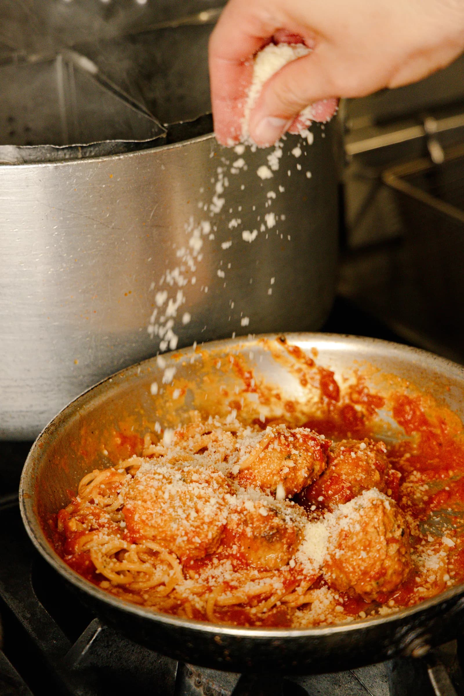A hand grating cheese over a pan of pasta and meatballs on the stove